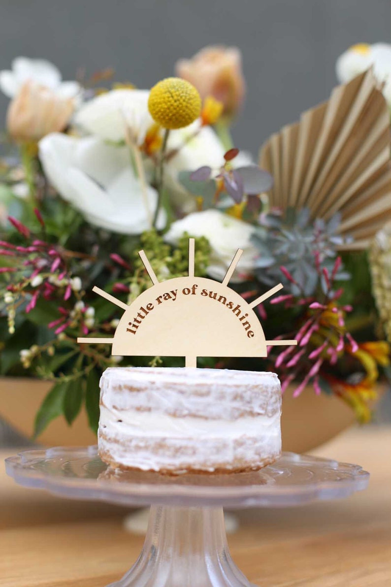 "little ray of sunshine" cake topper designed in the shape of a half sun with rays coming out. Topper is on a piece of cake, and there are flowers in the background. All Via Unmeasured Events