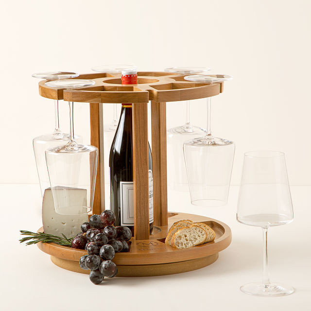 Wine and cheese wooden carousel that is monogrammed at the bottom. There are wine glasses hanging from the top and an assortment of fruit and cheese with a bottle of wine in the middle