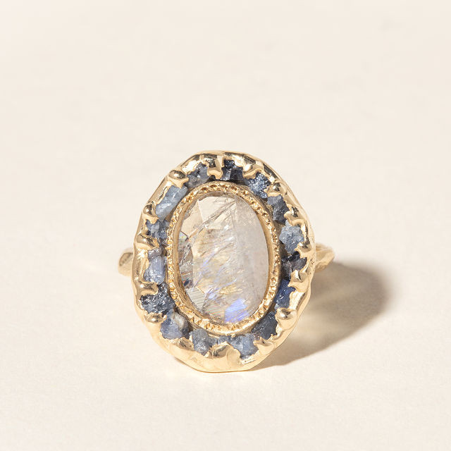 photo of a 14k lunar moonstone gem ring that has a spattering of blue moonstones around the large white moonstone. There is a gold band and the gem is facing the camera