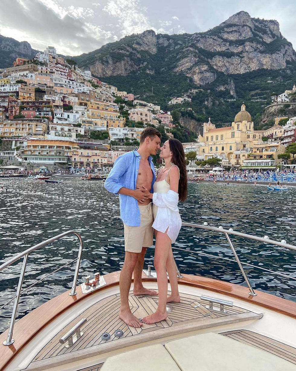 couple on a boat in the Amalfi coast of Italy as they look lovingly at one another. colorful building and mountains are seen in the background. all via unmeasured events