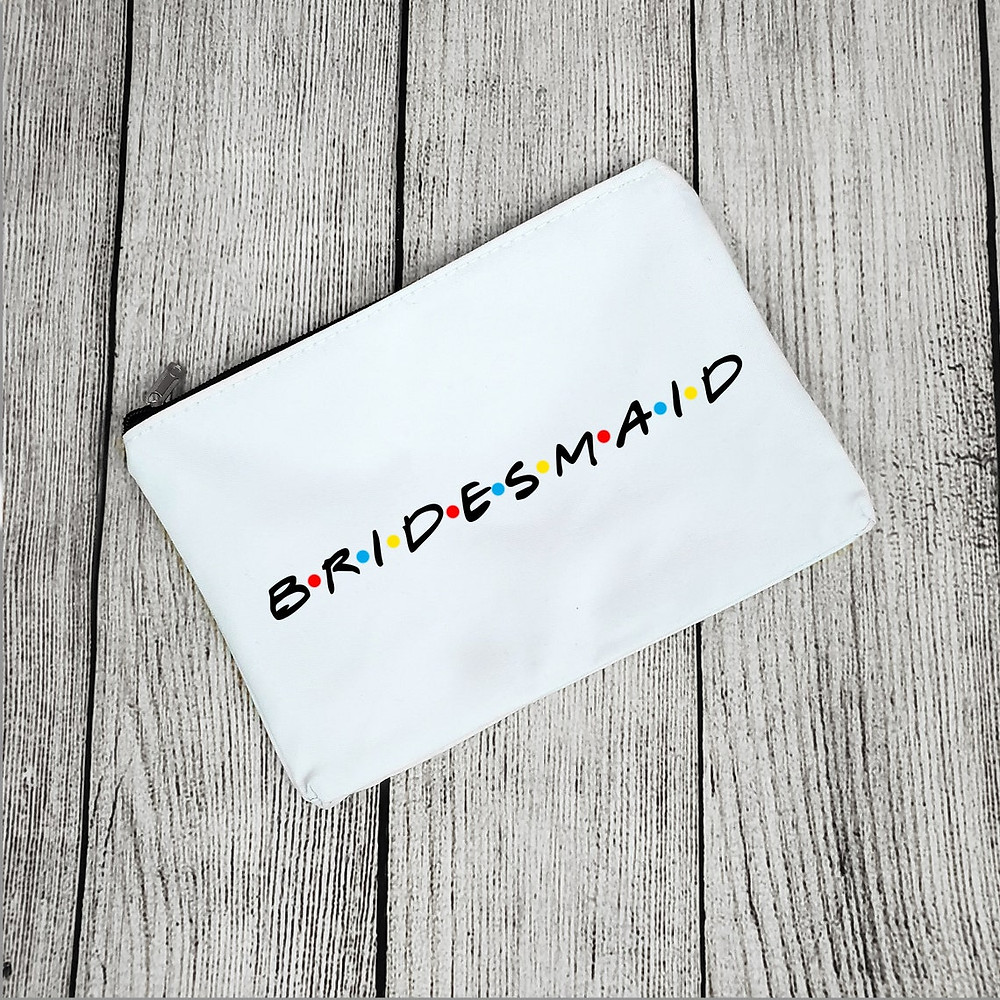 photo of a white makeup bag on a wooden surface that reads "BRIDESMAID" in the "Friends" themed lettering. Enlarged for detail. All Via Unmeasured Events
