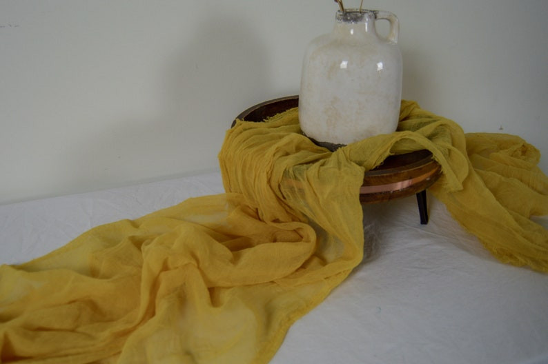 photo of a honey golden yellow colored cheesecloth table runner draped along a white floor and wooden table to show detail. A white ceramic vase sits on top. featured on the Unmeasured Events Blog