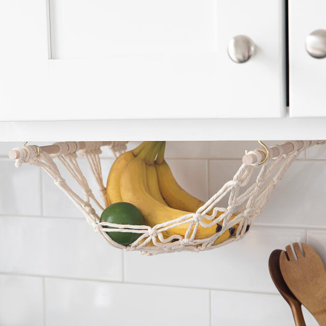 Banana and limes having from a macrame fruit hammock that is hanging from the bottom of a cupboard in a kitchen so fruit can hang