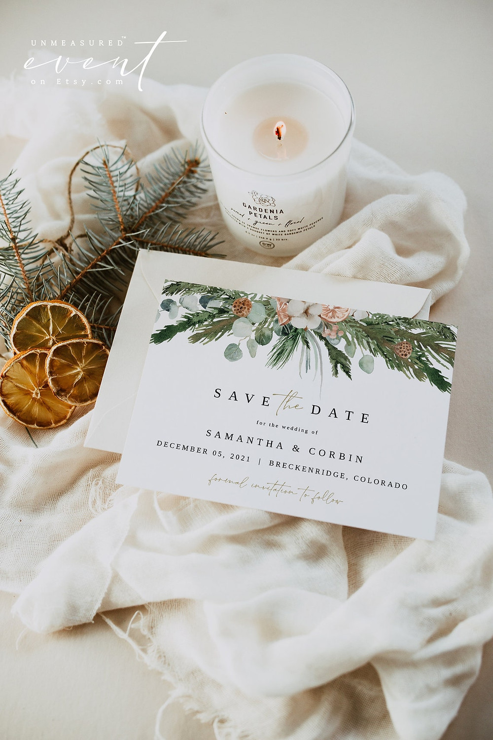 INGE collection Christmas template save the date from unmeasured events that shows Christmas pine trees and florals and energy handwritten font on a white card. postcard is placed on a white blanket with a pine tree, candle, and orange spice peels in the background. All via unmeasured events.