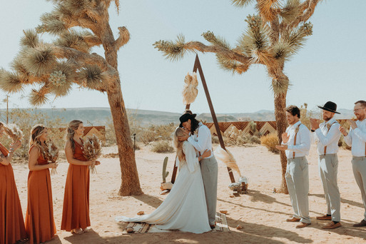 bride and groom kissing at their boho wedding alter with members of their bridal party on either side, and the desert in the background. All via Unmeasured Events