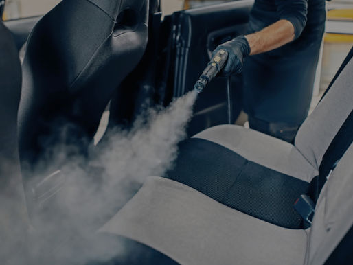 Person steam-cleaning a car seat with a black and gray fabric in a garage. The gloved hand holds a nozzle emitting steam, creating a focused mood.