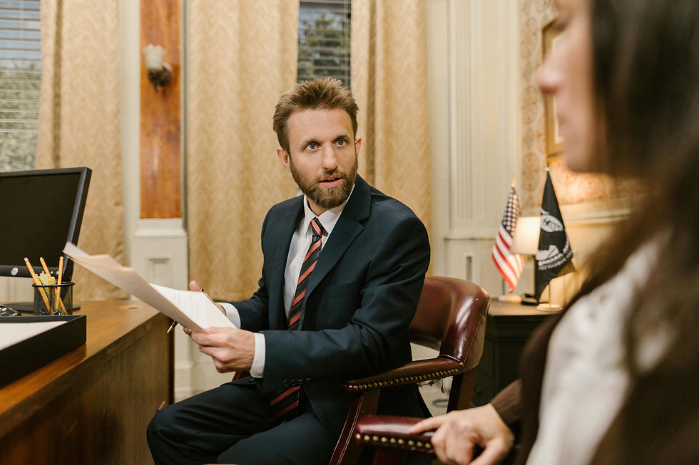 Man in suit holding papers, sitting at a desk facing a lawyer with computer. Engaged in a serious conversation with a woman.