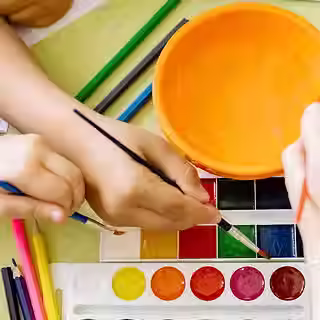 Hands holding paintbrushes above a set of watercolor paints.