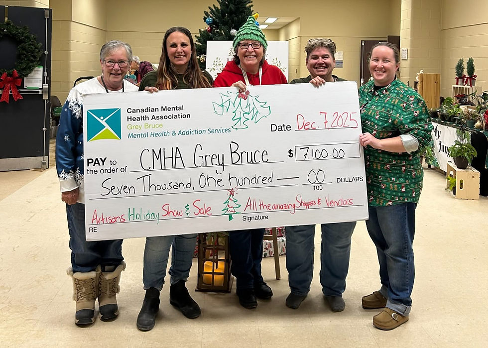 A group of five Artisans Holiday Show & Sale committee members stand together holding an oversized cheque for CMHA Grey Bruce. The cheque displays a donation amount of $7,100 from the 2025 Artisans Holiday Show & Sale. Holiday decorations and artisan booths are visible in the background.
