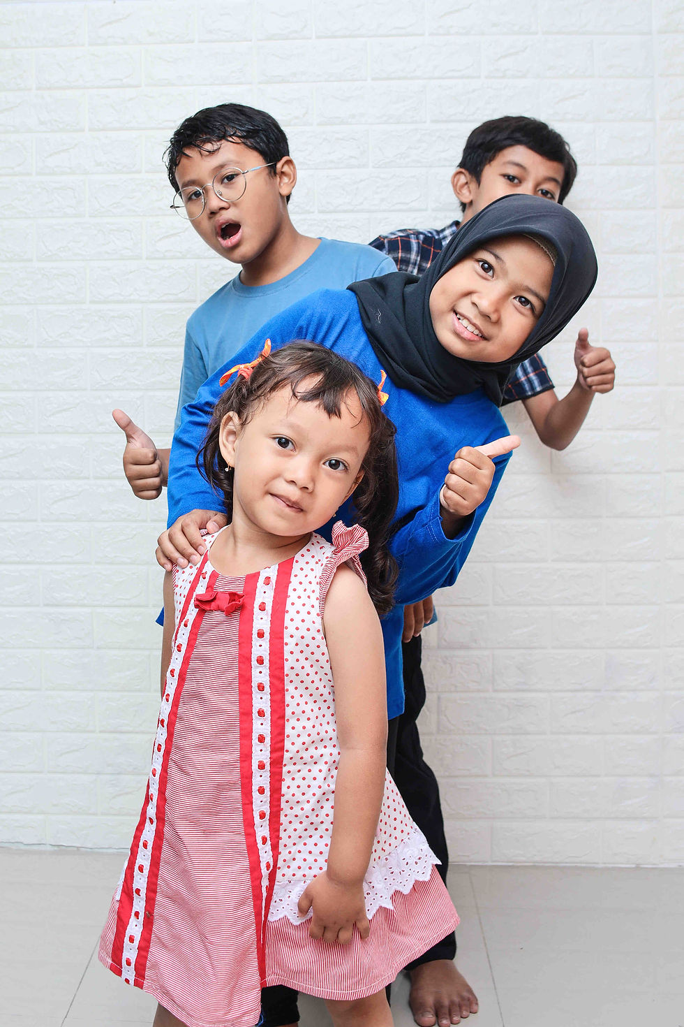 Four smiling kids pose for a photo with thumbs up near a white wall.