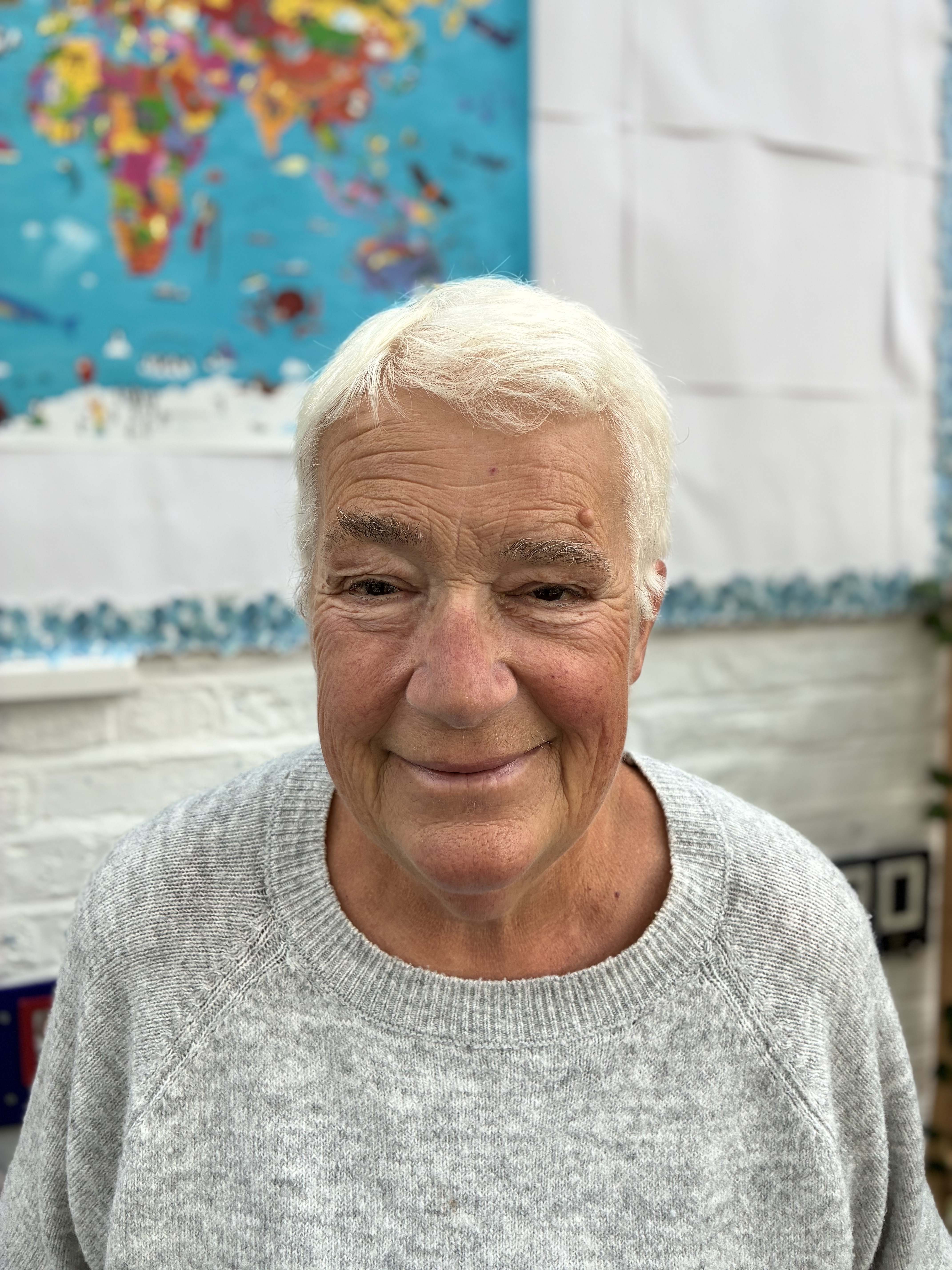 Smiling older woman in grey shirt poses next to a world map.