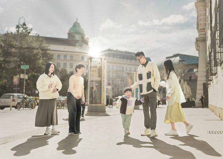 A family of five, including three adults and two children, engages in an elegant family photoshoot on a sunny day along a Vienna street, featuring historic buildings like Albertina and Opera in the background.