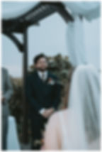A groom in a dark suit smiles at a bride under an outdoor arch with white drapes, surrounded by foliage, photographed by Blackout Photography.