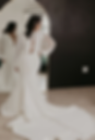 Woman in a long white bridal gown stands before a full-length mirror in a minimalist room, her train spread on the floor.