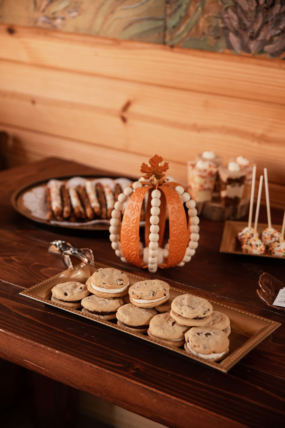 Wooden table with cookies, cake pops, and parfaits. Decorative orange pumpkin centerpiece. Warm, rustic setting with neutral tones.