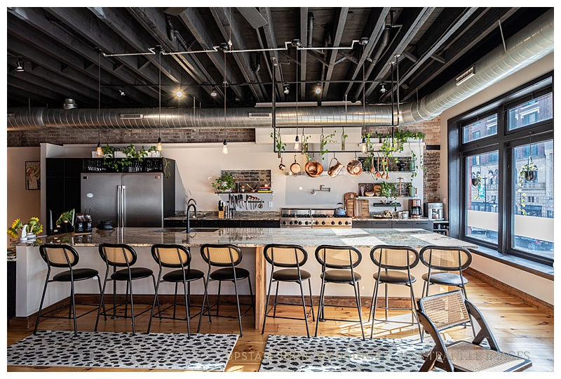 Modern kitchen with eight high chairs at a marble island, hanging copper pots, plants, light wood floor, large windows, and exposed beams.