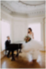 Bride in white gown holds bouquet beside a man playing piano in an ornate, sunlit room. Elegant, romantic ambiance.