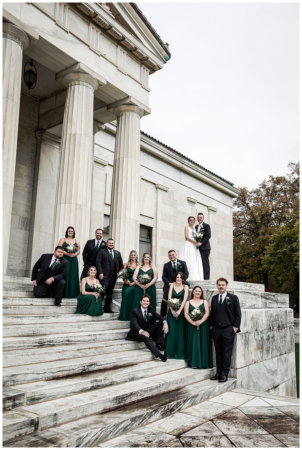 Wedding party on marble steps, with bridesmaids in green dresses and groomsmen in dark suits. A couple stands smiling, holding flowers. Classic building backdrop.