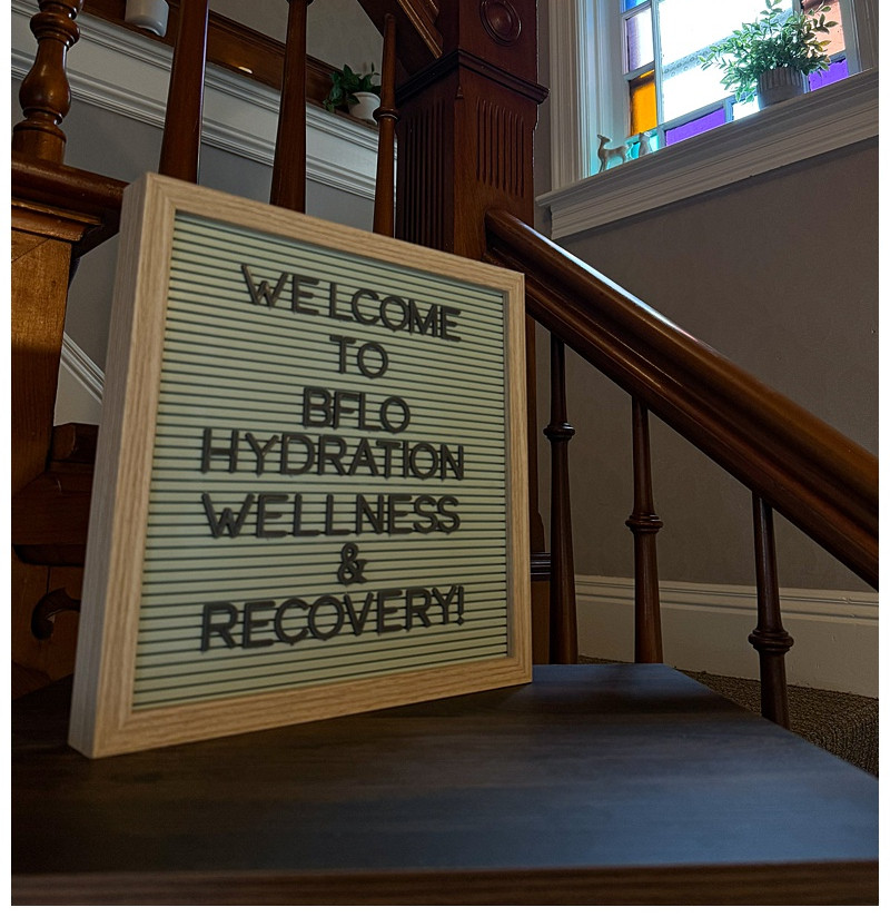 Framed sign on a wooden staircase reads Welcome to Bflo Hydration Wellness & Recovery beside a colorful stained-glass window.