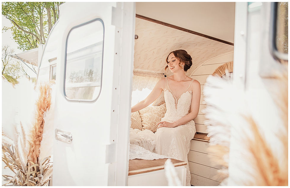 Bride in a lace dress sits in a white vintage trailer, smiling. Soft lighting, pampas grass, green trees visible through window.