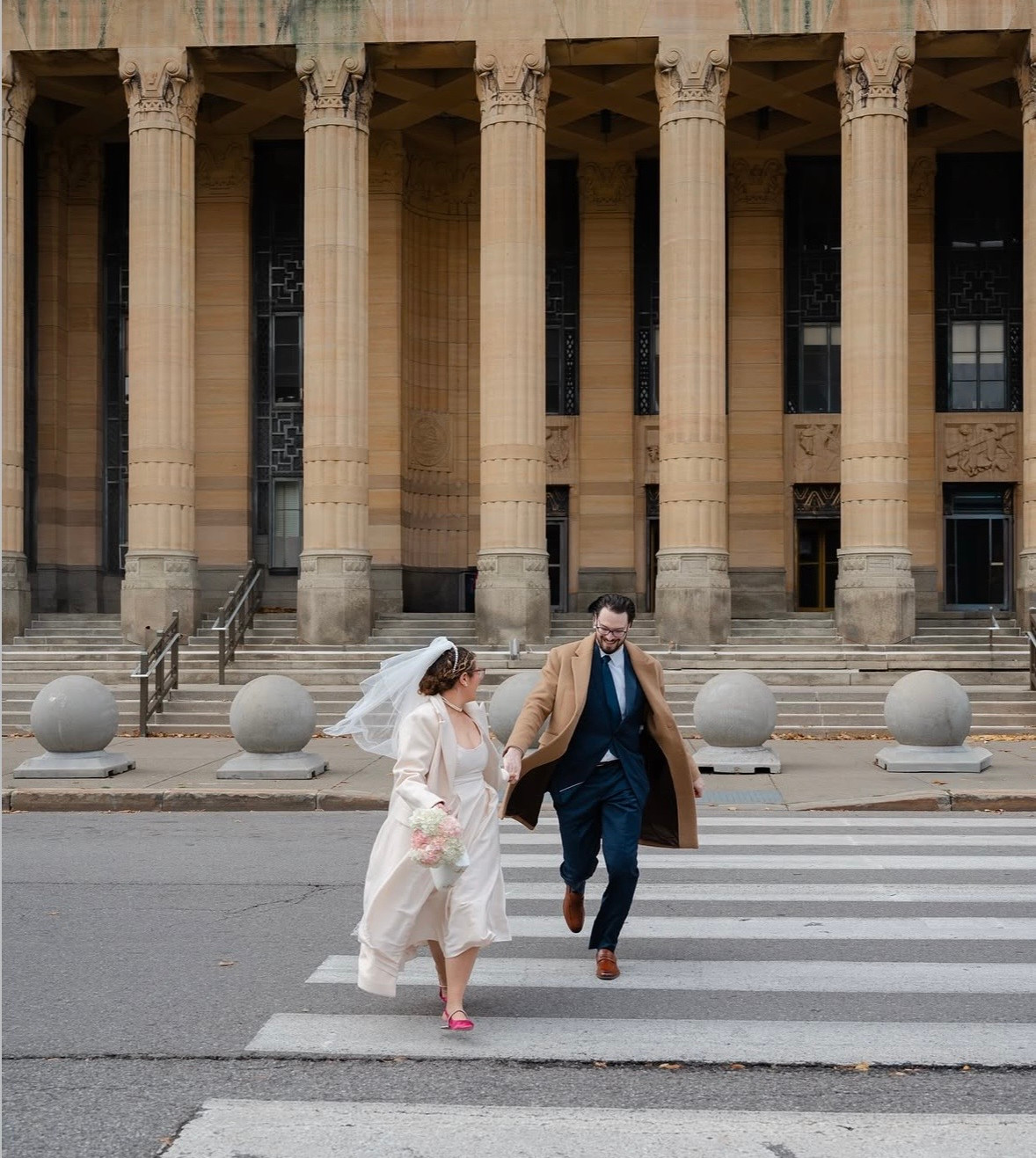 Bride and groom run hand in hand across a crosswalk in front of a grand stone building with tall columns, wny wedding vendors, wny wedding inspiration, wny wedding ideas, wny photographers