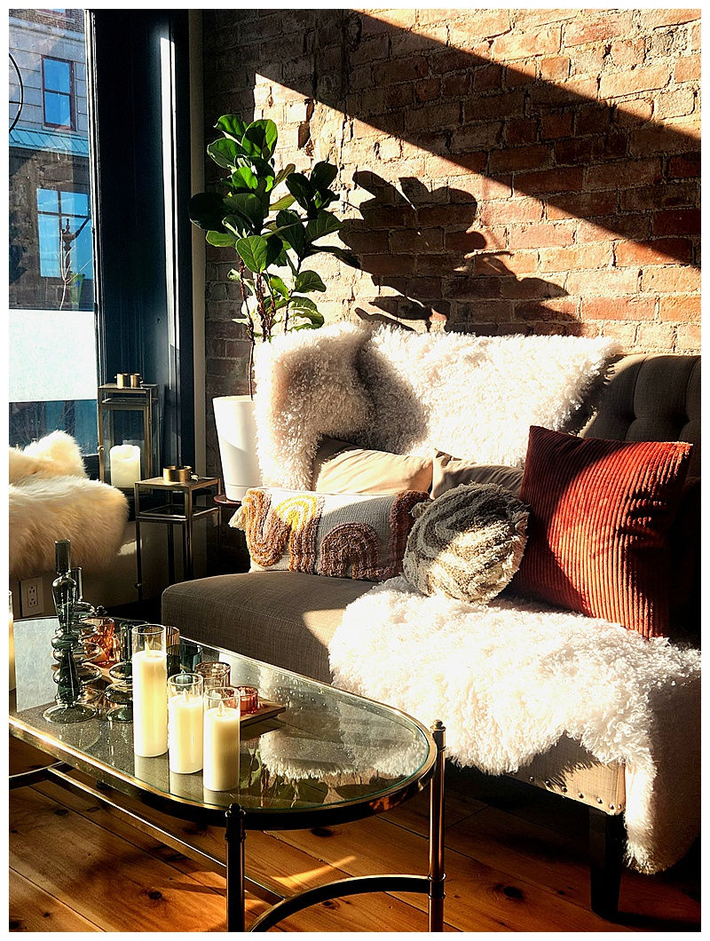 Cozy living room with sunlight casting shadows on brick wall. Sofa with textured pillows, fluffy throws. Glass table with candles, plant nearby.