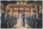 Bride and groom kiss in a grand church aisle, bride in white gown with bouquet, groom in blue suit, ornate marble and gold decor.
