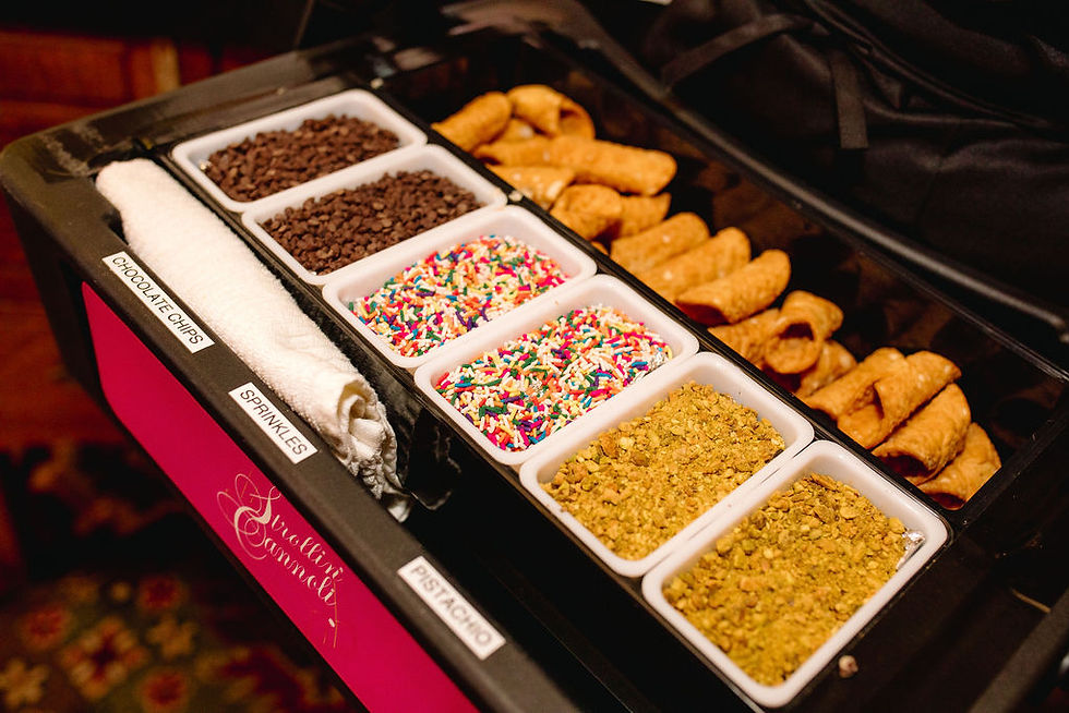 Cannoli toppings bar with trays of chocolate chips, sprinkles, and pistachios. Rolled cannoli shells are in the background.