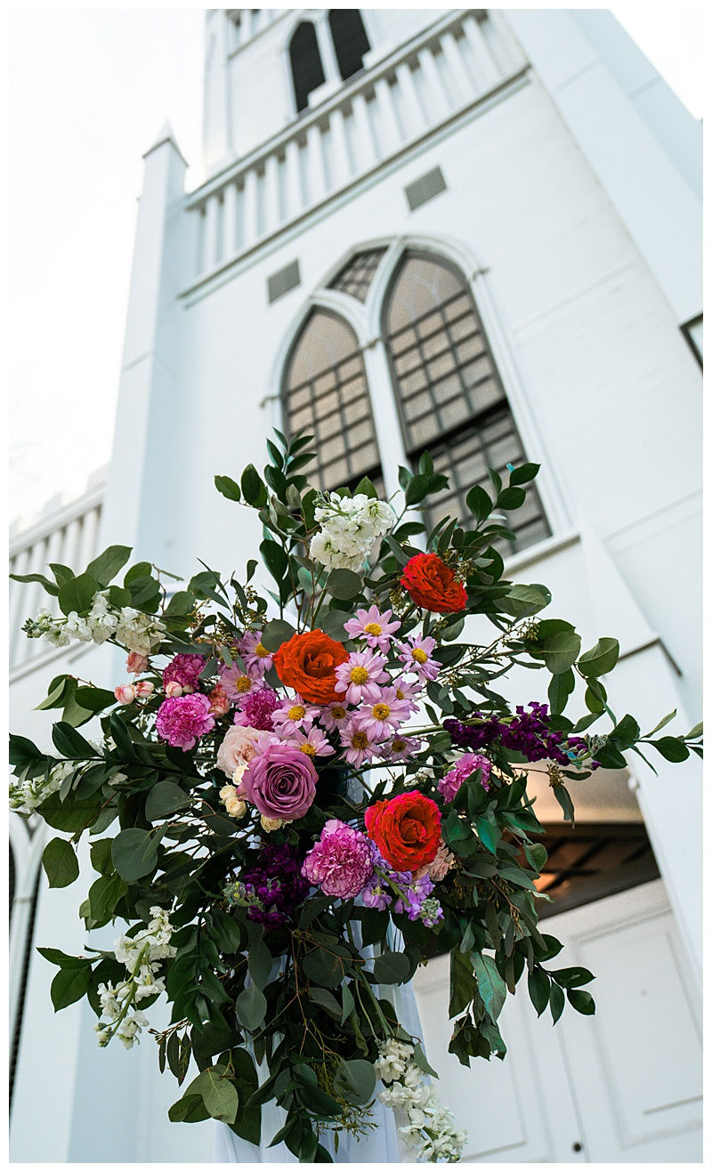 Bouquet of bright red, pink, and purple flowers with greenery against a white building with arched windows, photographed by Blackout Photography.