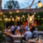 Outdoor patio dinner party with glowing string lights above blurred guests seated at tables at dusk