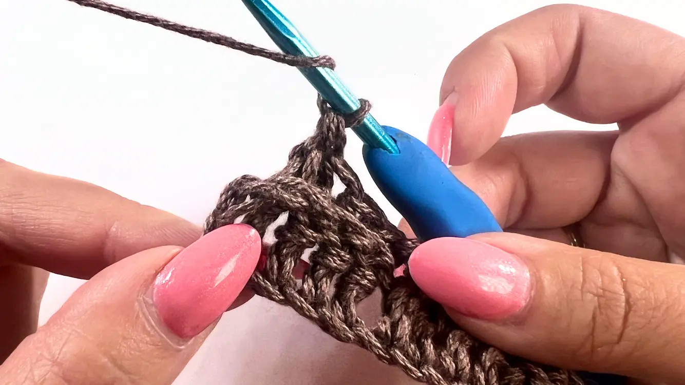 Close-up of a regular double crochet (DC) being made into the top of the turning chain at the end of the row, shown with a blue crochet hook and brown yarn.