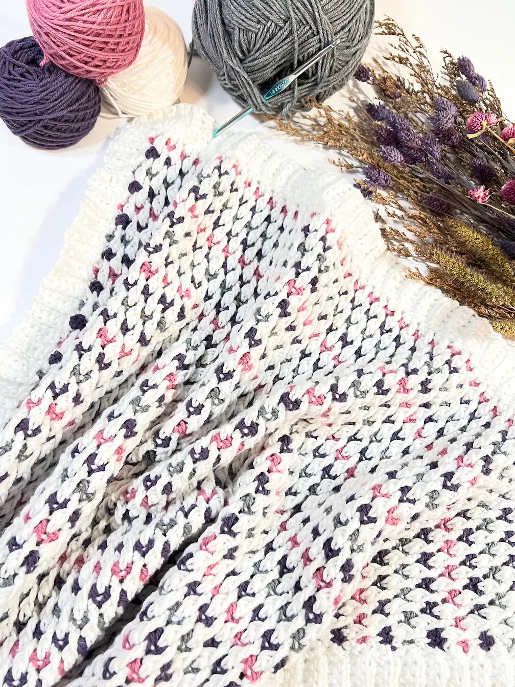 Flat lay of the Cuddle Blanket crochet pattern with soft white, pink, grey, and purple yarns beside a crochet hook and dried flowers.