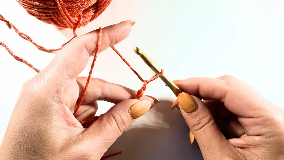 Hands demonstrating how to hold crochet yarn, hook, and project together while making foundation chain stitches with rust-coloured yarn.