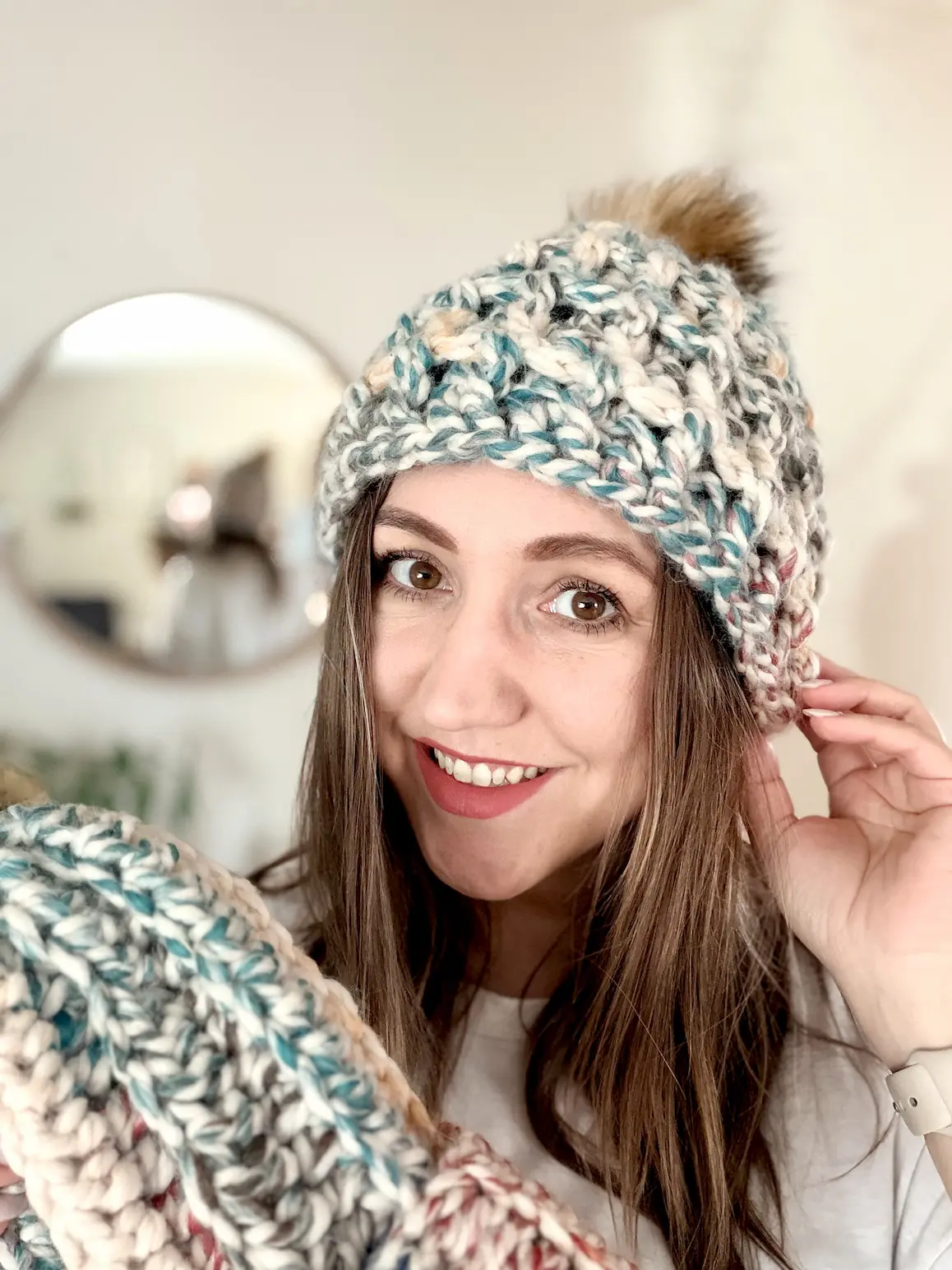 Woman smiling while wearing chunky crochet beanie with faux fur pompom, made from super bulky variegated yarn in neutral tones.