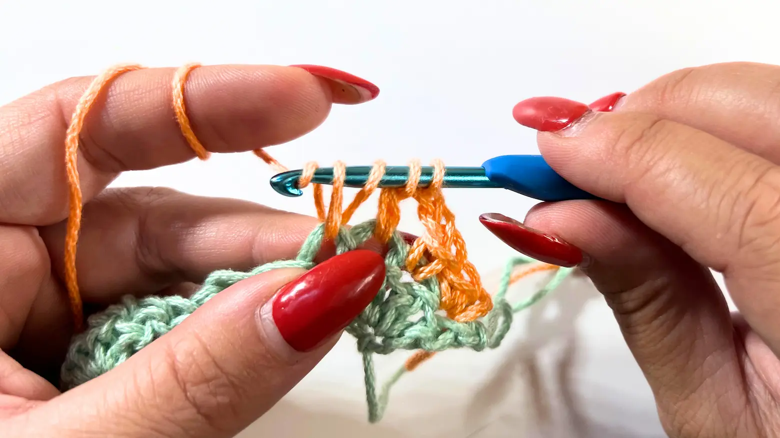 Close-up of Row 2 in progress with orange yarn. The blue crochet hook holds four loops after working several partial double crochets for the 7DC together cluster.