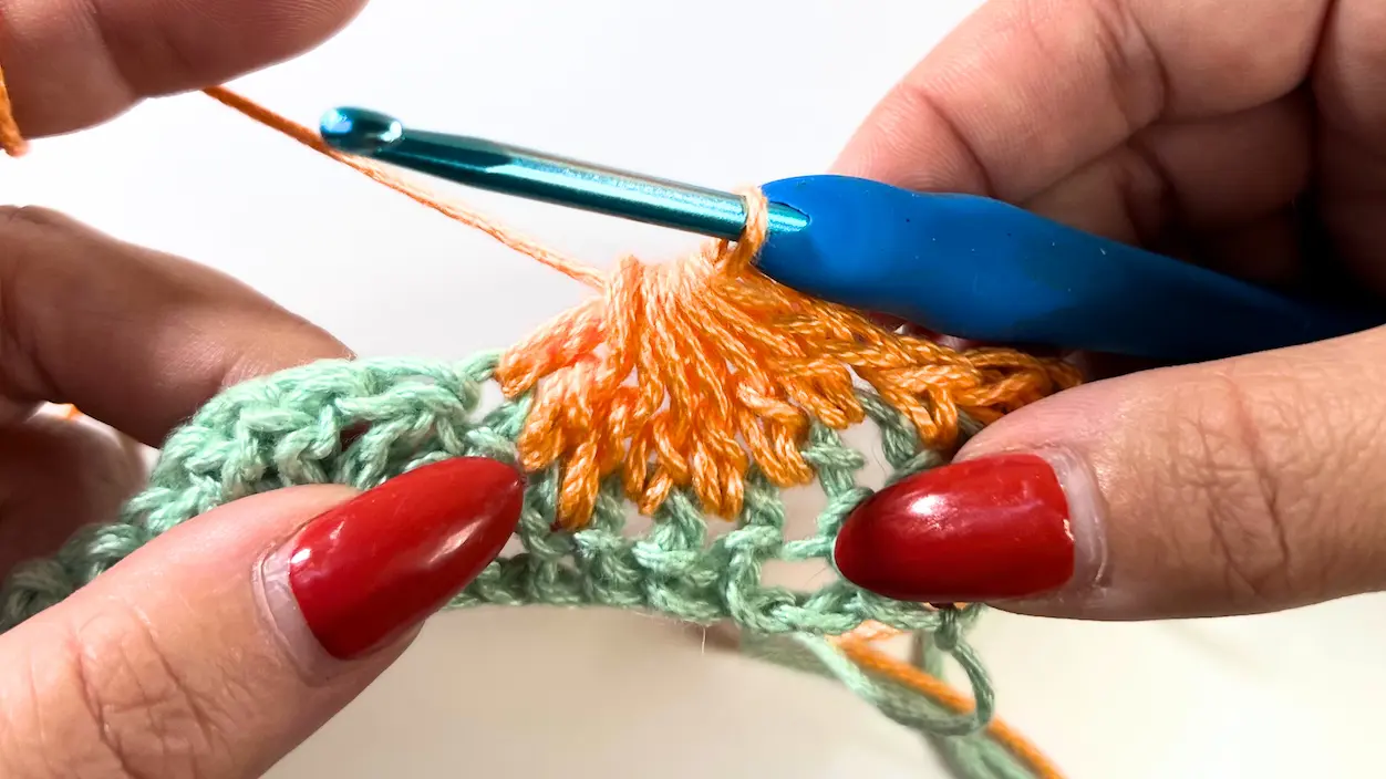 Row 2 of the Catherine Wheel Stitch in orange yarn. The blue crochet hook has just pulled through all 8 loops on the hook, completing the 7DC together cluster, which forms a closed fan shape over the green row.