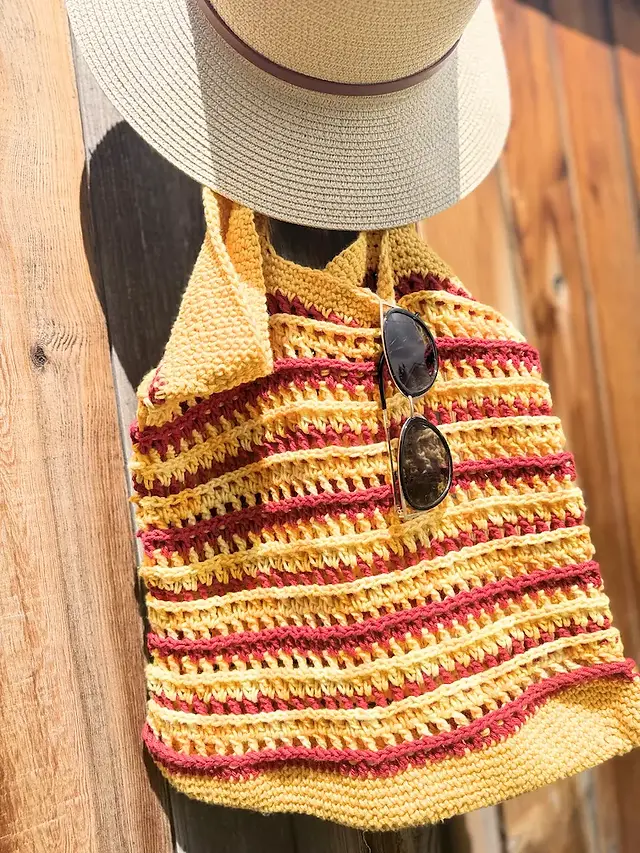 Finished crochet summer beach tote bag with textured striped pattern in yellow and burgundy yarn, hanging on a wooden fence with a sun hat and sunglasses.