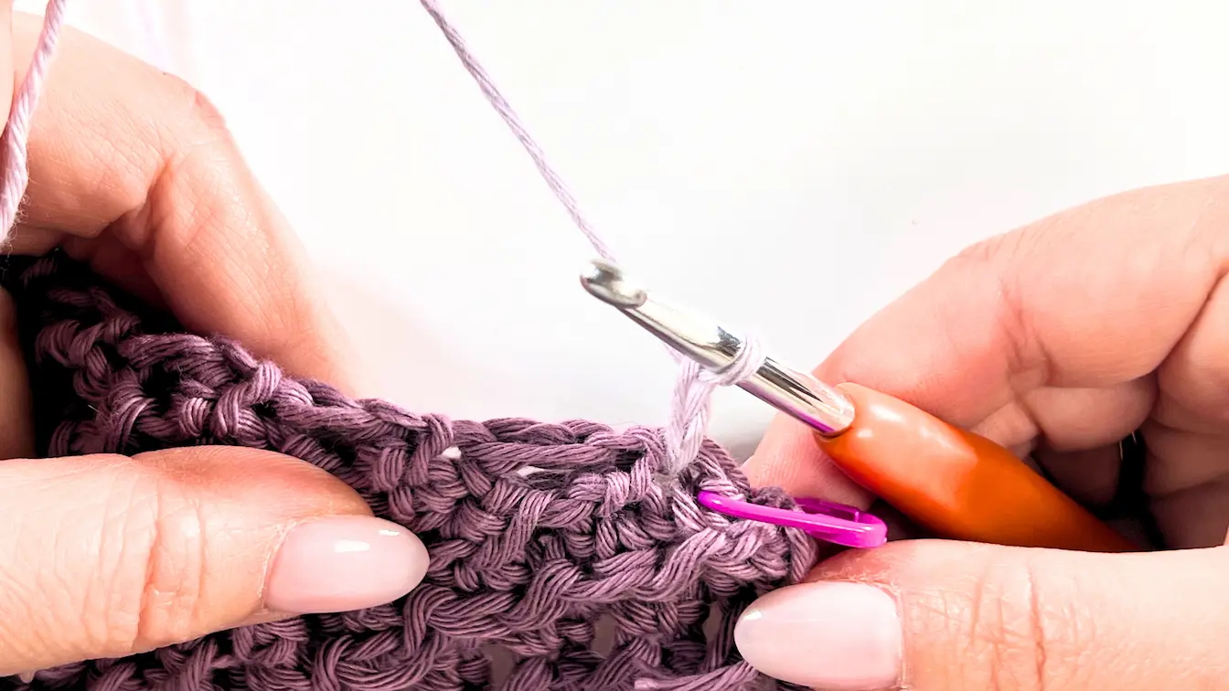 Close-up of a crochet hook inserted through both layers of the hood and towel at a marked point using a pink stitch marker. The working yarn is looped on the hook, ready to begin the seaming process in dusty lavender cotton yarn.