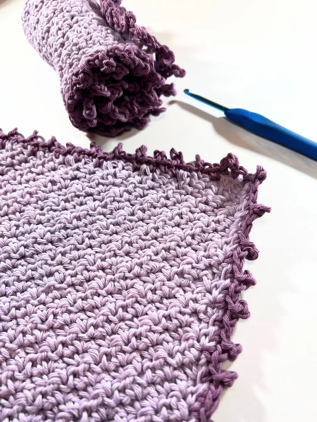 Rolled and flat crochet washcloths with a textured lemon peel stitch pattern and purple picot border, shown with a blue crochet hook on the side.