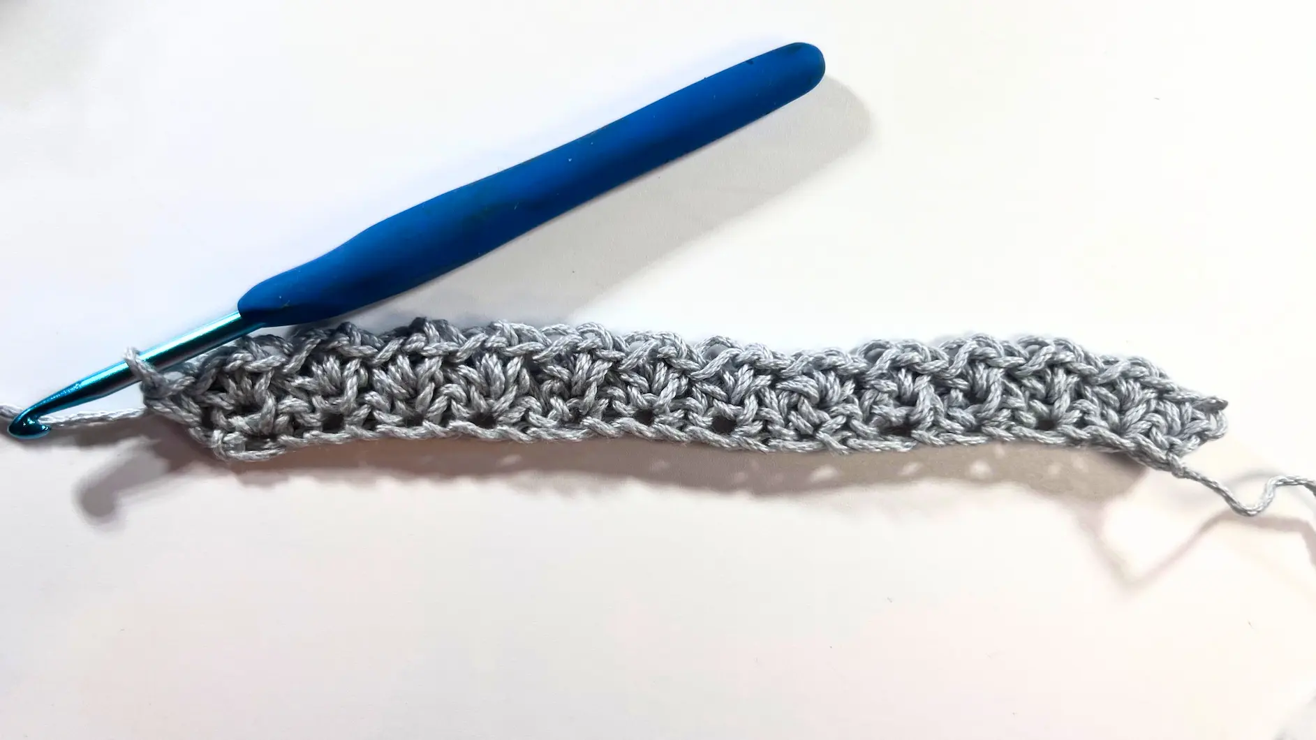 Full view of the crochet swatch after completing two rows of Spider Stitch. A blue-handled hook is attached to the final stitch, with visible texture forming across the grey yarn.