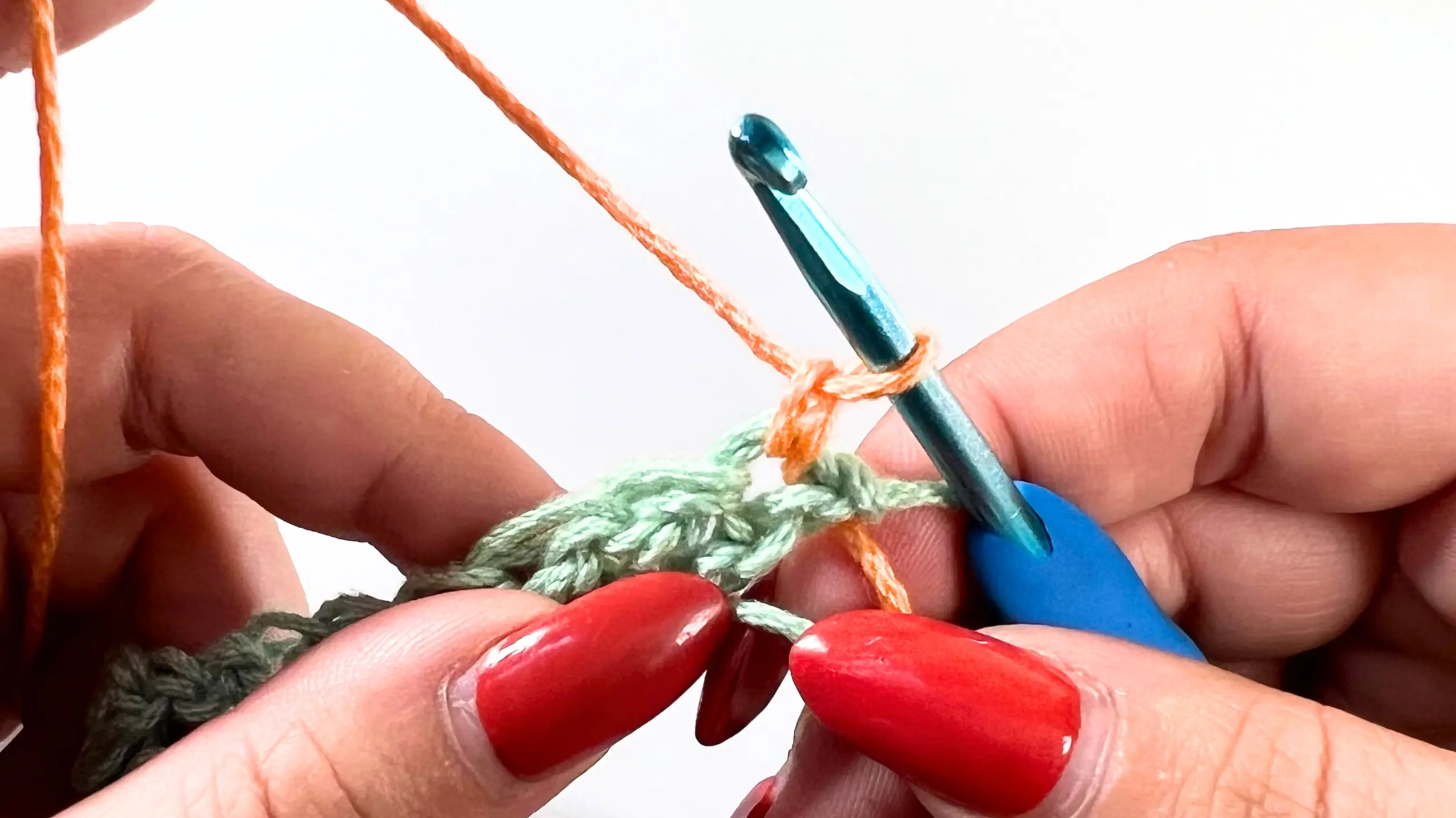 Close-up of the start of Row 2, with orange yarn secured by a chain stitch on the blue crochet hook. The green stitches from Row 1 are visible underneath.