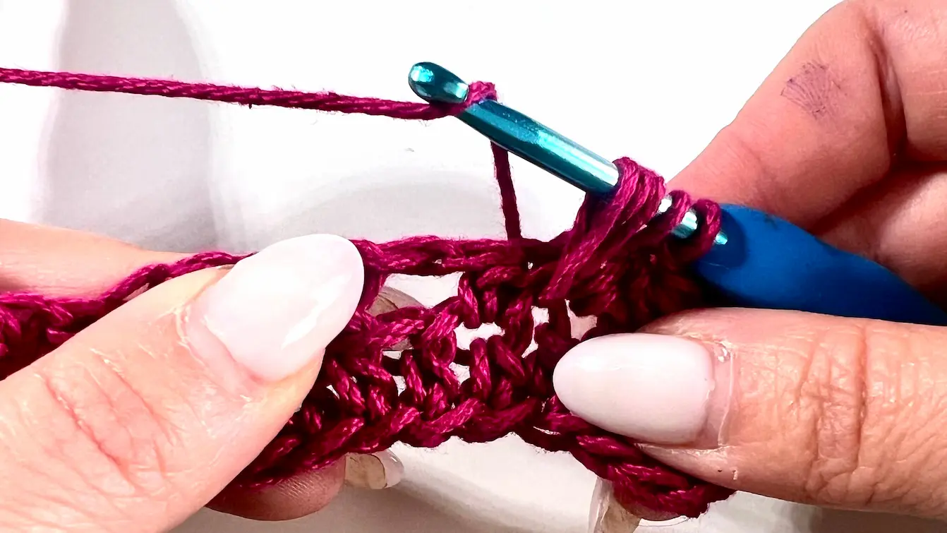 Close-up showing how to crochet the feather stitch step-by-step using fuchsia yarn and a blue crochet hook — part of a textured stitch tutorial.