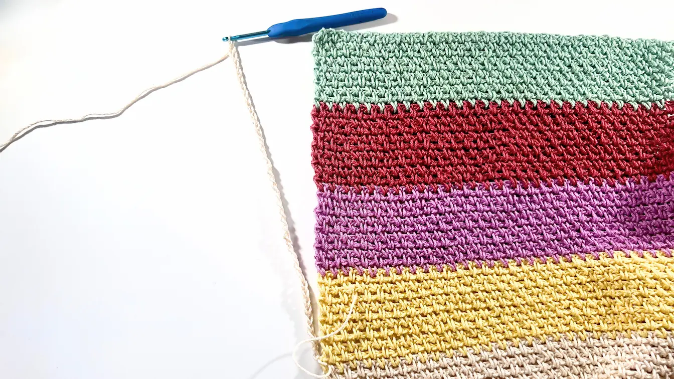 A colourful crochet panel laid flat with a new foundation chain extending from the edge in a new yarn colour, marking the beginning of the second half of the panel.