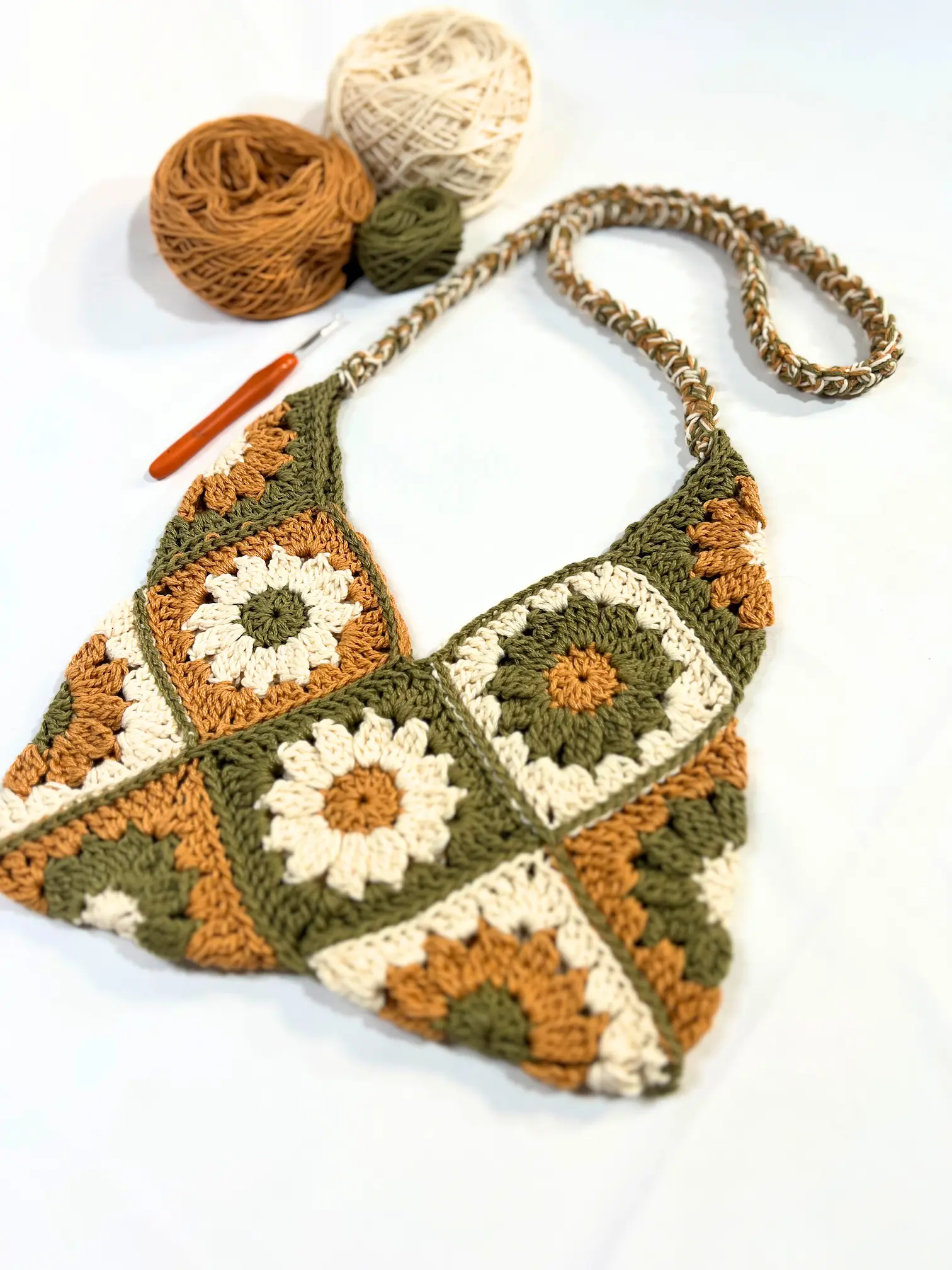  Finished crochet Meadow Daisy Bag laid flat, with a crochet hook and matching yarn balls in earthy cream, green, and copper colours displayed beside it.