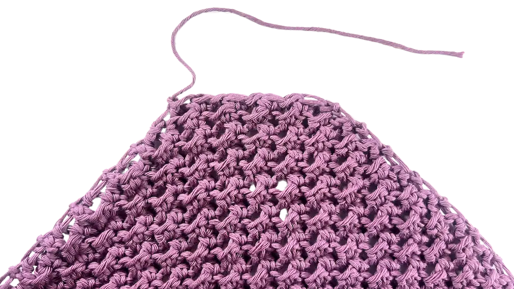 Triangular crochet piece created using Corner-to-Corner Even Moss Stitch in dusty lavender cotton yarn. The final decrease rows are complete, and a long yarn tail is visible for seaming or edging.