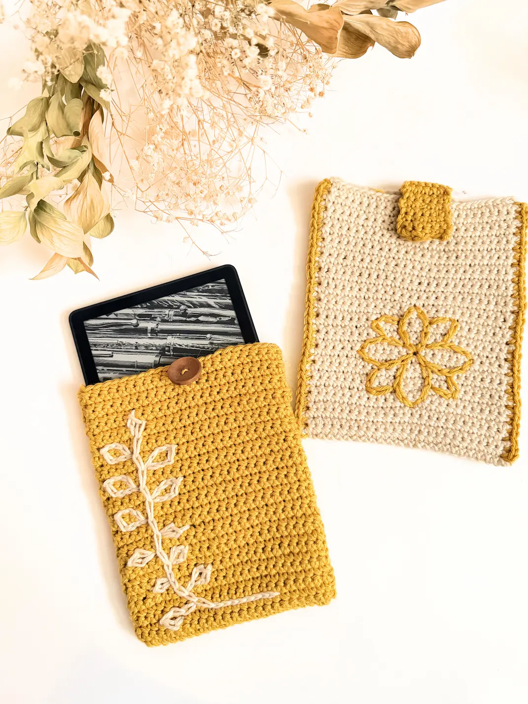 Mustard crochet Kindle sleeve with a white leaf surface crochet design and wooden button closure, displayed with a Kindle e-reader.