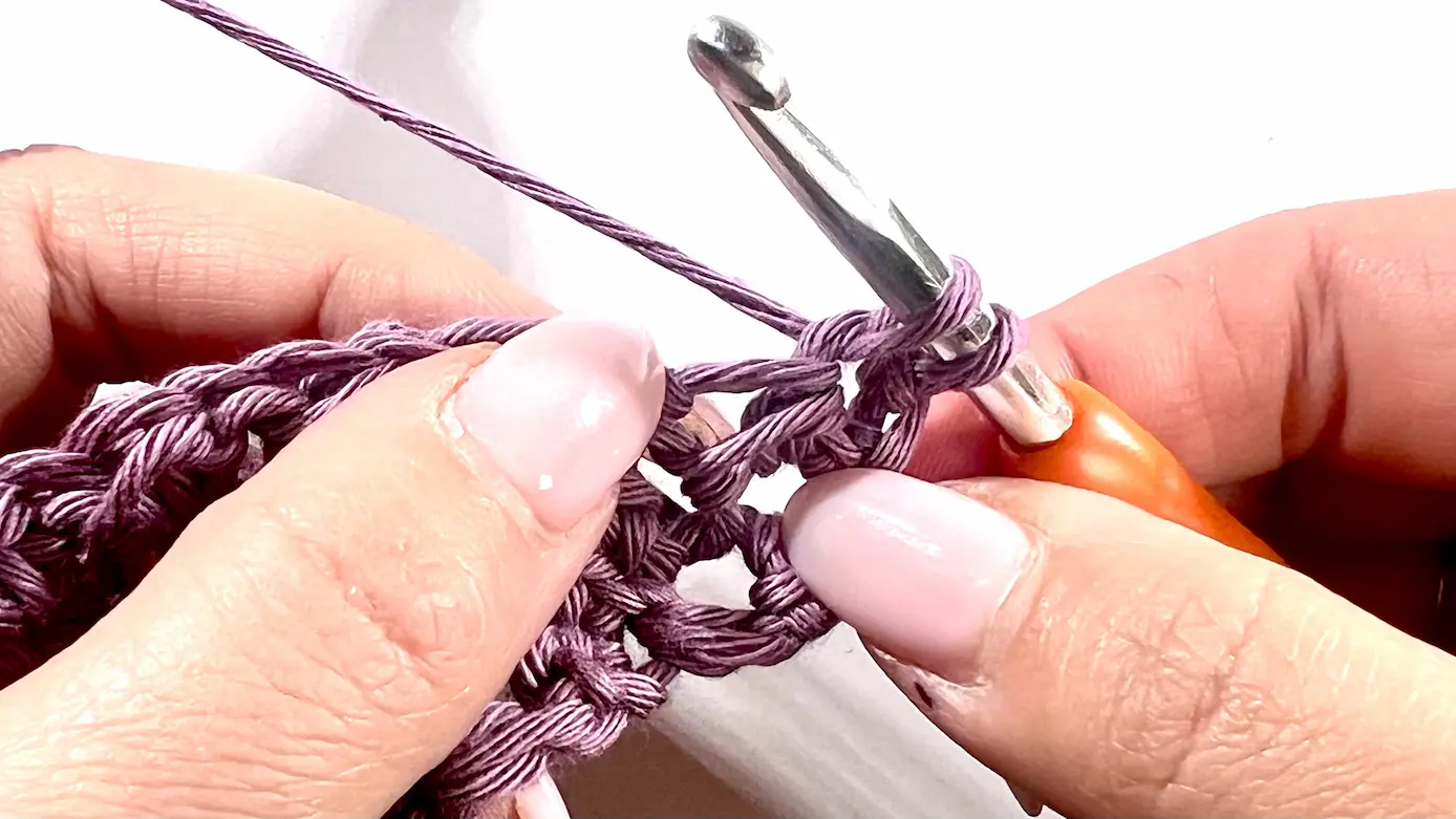 Crochet hook being inserted into the second stitch space as part of the corner-to-corner decrease in Even Moss Stitch. Light purple yarn and a textured stitch pattern are clearly visible.