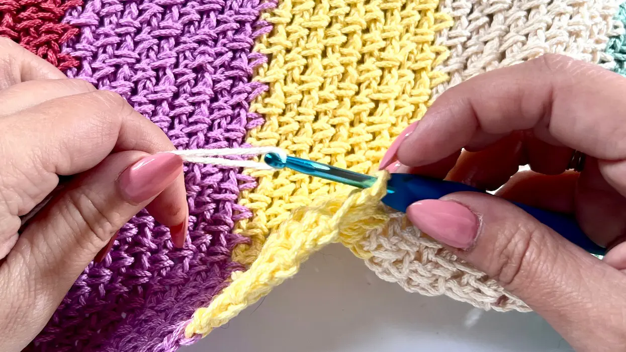 Hands holding a crochet hook and yarn while attaching a new colour (cream) to the edge of a striped crochet panel, just before pulling through to join.