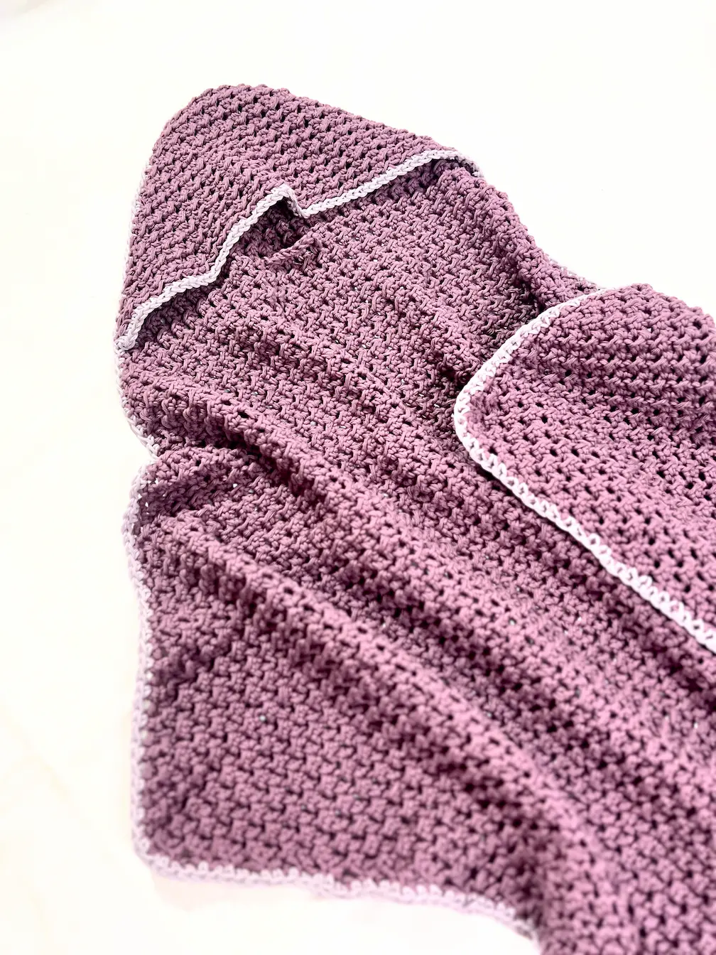 Handmade crochet baby towel in dusty lavender yarn with a contrasting light grey border, hanging neatly from the hood. The hood is folded over the top, showing off the corner-to-corner Even Moss Stitch texture.