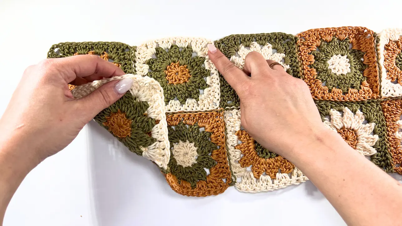 Hands holding a crochet daisy square section, beginning the first fold by lifting one corner towards the center.